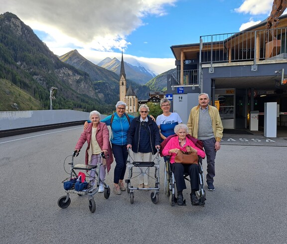 Sechs Senior*innen vor Bergkulisse und Kirche in Heiligenblut, zwei mit Rollator, eine im Rollstuhl