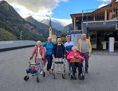 Sechs Senior*innen vor Bergkulisse und Kirche in Heiligenblut, zwei mit Rollator, eine im Rollstuhl