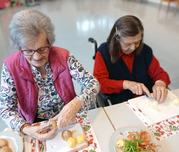 Zwei Seniorinnen sitzen am Tisch und schneiden Kartoffeln und Zwiebeln für das Essen.
