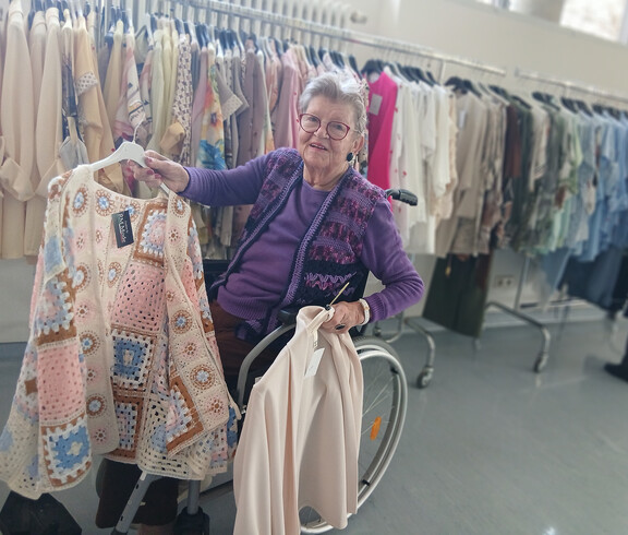 Bewohnerin im Rollstuhl hält helle Kleidung vor einer Reihe bunter Oberteile