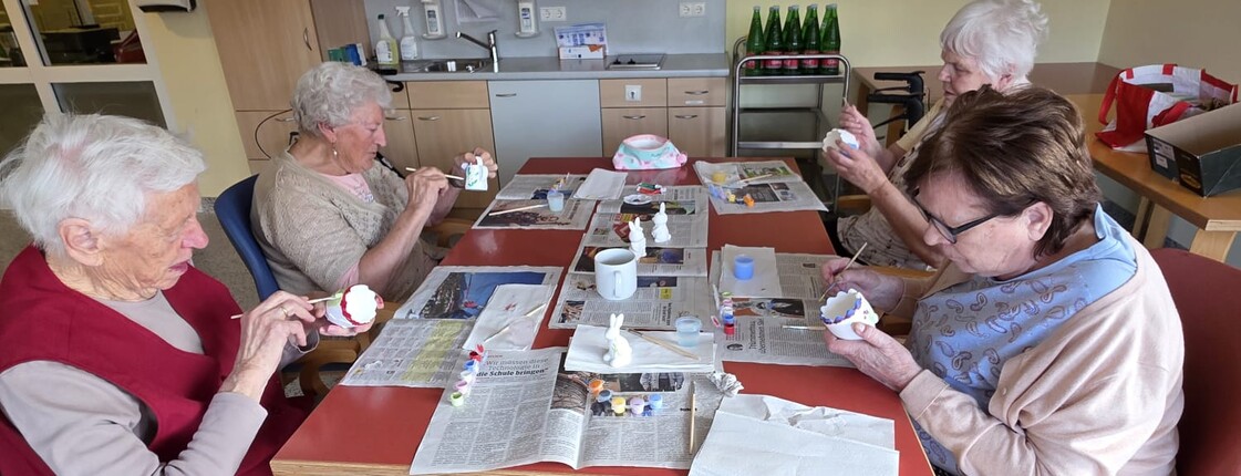 Bewohnerinnen des Pflegewohnhauses bemalen gemeinsam Keramikfiguren und gestalten farbenfrohe Osterdekorationen an einem Arbeitstisch.