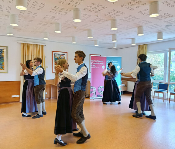 Mehrere junge Paare tanzen Volkstänze in Tracht vor älteren Zuschauer*innen