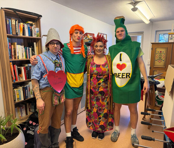 Vier verkleidete Mitarbeitende posieren gemeinsam in einem Zimmer mit Bücherregal und Deko.