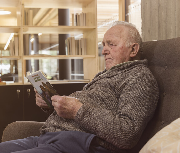 Das Bild zeigt eine Person im Tageszentrum, die in einem ruhigen, gemütlichen Aufenthaltsbereich sitzt und ein Buch liest, umgeben von Holzregalen und einer wohnlichen Atmosphäre.