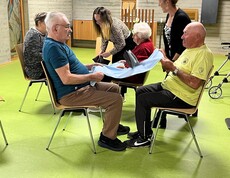 Bewohner*innen des Pflegewohnhauses sitzen sich paarweise gegenüber und führen mit Unterstützung von Mitarbeitenden eine Bewegungsübung mit einem Tuch zur Förderung von Mobilität und Koordination durch.