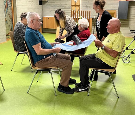Bewohner*innen des Pflegewohnhauses sitzen sich paarweise gegenüber und führen mit Unterstützung von Mitarbeitenden eine Bewegungsübung mit einem Tuch zur Förderung von Mobilität und Koordination durch.