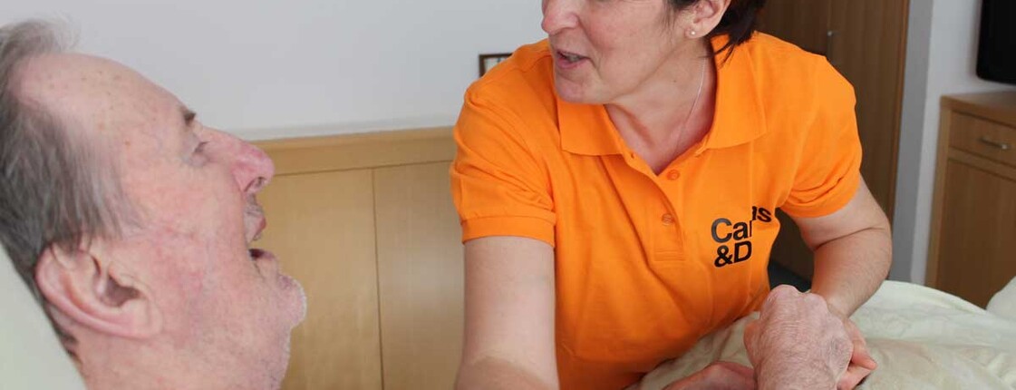 Ein Mann liegt im Bett und eine Caritas-Mitarbeiterin hilft ihm die Arme zu stecken.
