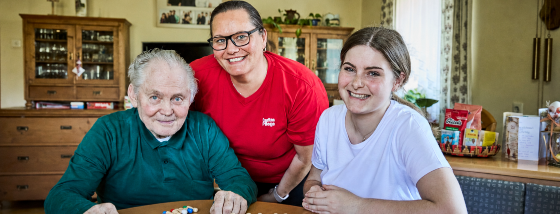 Pflegefachkraft und Schülerin sind zu Hause bei einem Kunden der mobilen Pflege. Sie sitzen mit ihm an einem Tisch uns spielen ein Brettspiel.