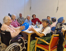Senior*innen am Tisch, eine Frau im Rollstuhl spielt lachend mit einem Ballon, festliche Stimmung