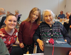 Bewohnerin hält ein schwarzes Geschenk mit roter Schleife, daneben eine junge Dame und en Mädchen.