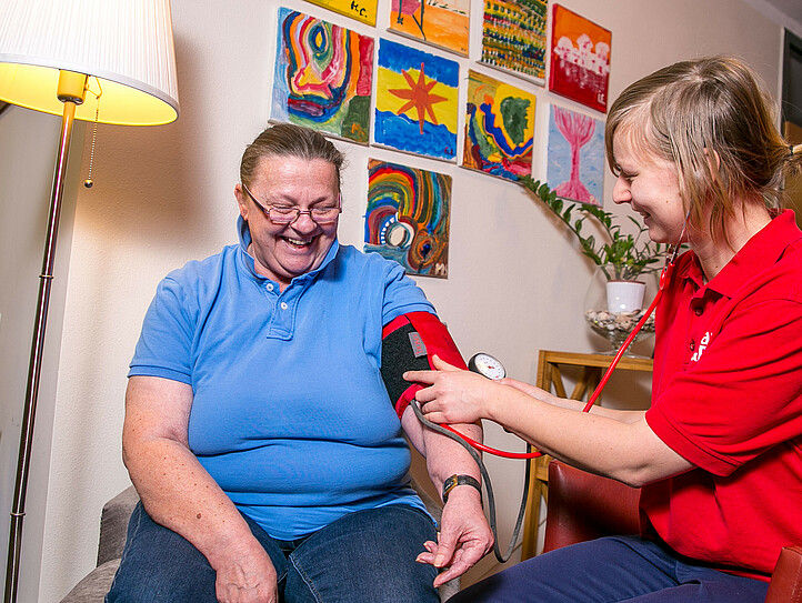 Eine Caritas-Pflegerinmisst den Blutdruck bei einer Frau, die beiden lachen.