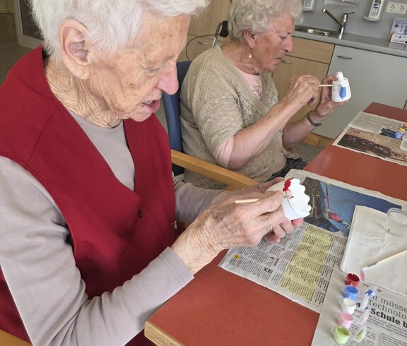 Im Gemeinschaftsraum des Pflegewohnhauses sitzen zwei Bewohnerinnen an einem Tisch und bemalen konzentriert kleine Keramikfiguren. Auf dem Tisch liegen Zeitungen, Pinsel und Farbtöpfchen, die für die kreative Gestaltung genutzt werden.