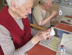 Im Gemeinschaftsraum des Pflegewohnhauses sitzen zwei Bewohnerinnen an einem Tisch und bemalen konzentriert kleine Keramikfiguren. Auf dem Tisch liegen Zeitungen, Pinsel und Farbtöpfchen, die für die kreative Gestaltung genutzt werden.