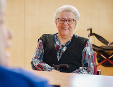 Seniorin sitzt mit Gehstock im Aufenthaltsraum während eines Gesprächs.