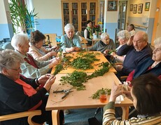 Bewohnerinnen und Bewohner des Pflegewohnhauses sitzen im Gemeinschaftsraum zusammen und binden in fröhlicher Runde Palmbuschen. Auf dem großen Tisch liegen frische Zweige bereit, während gemeinsam gearbeitet, erzählt und gelacht wird.
