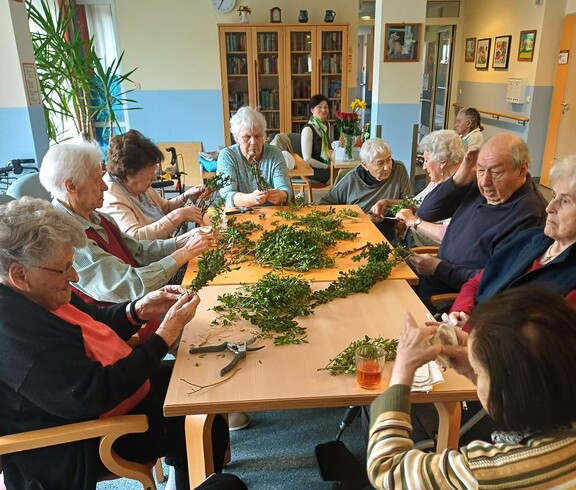 Bewohnerinnen und Bewohner des Pflegewohnhauses sitzen im Gemeinschaftsraum zusammen und binden in fröhlicher Runde Palmbuschen. Auf dem großen Tisch liegen frische Zweige bereit, während gemeinsam gearbeitet, erzählt und gelacht wird.