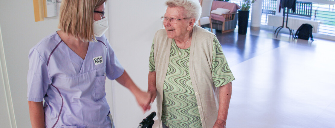 Eine Kollegin aus der Caritas Pflege im Pflegewohnhaus im Gespräch mit einer älteren Dame mit Rollator.