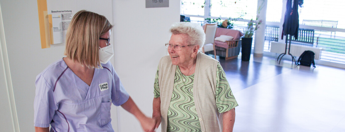 Eine Kollegin aus der Caritas Pflege im Pflegewohnhaus im Gespräch mit einer älteren Dame mit Rollator.