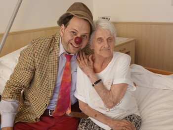 Frau sitzt mit Clown im Bett beide lächeln in die Kamera