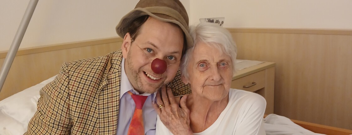 Frau sitzt mit Clown im Bett beide lächeln in die Kamera