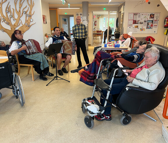 Eine Gruppe von Menschen sitzt in einem Gemeinschaftsraum. Zwei Personen spielen Akkordeon, während andere in Stühlen und Rollstühlen sitzen. 