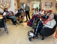 Eine Gruppe von Menschen sitzt in einem Gemeinschaftsraum. Zwei Personen spielen Akkordeon, während andere in Stühlen und Rollstühlen sitzen. 