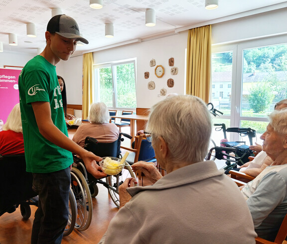 Jugendlicher verteilt Eis an mehrere Seniorinnen im Rollstuhl im Aufenthaltsraum