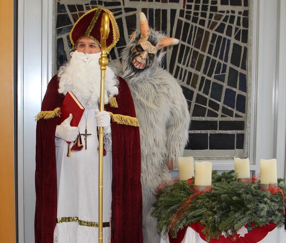 Nikolaus im roten Mantel mit goldenem Stab und rotem Buch steht neben Krampus mit hellgrauem Fell. Beide befinden sich im festlich geschmückten Pflegewohnhaus neben einem Adventkranz.