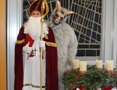 Nikolaus im roten Mantel mit goldenem Stab und rotem Buch steht neben Krampus mit hellgrauem Fell. Beide befinden sich im festlich geschmückten Pflegewohnhaus neben einem Adventkranz.