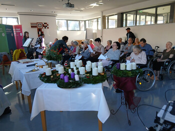 Mehrere Adventkränze mit Kerzen auf Tisch, Menschen sitzen dahinter und lesen