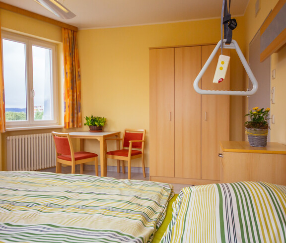 In einem hellen Zimmer stehen ein Tisch und zwei Stühle, Nachtkästchen, Kleiderschrank, Fenster, orange Vorhänge, Heizkörper und ein Bett für pflegebedürftige Personen mit frisch überzogener gelb-grün gestreifter Bettwäsche. Auf dem Tisch und dem Nachtkästchen befindet sich jeweils eine Zimmerpflanze. 
