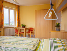 In einem hellen Zimmer stehen ein Tisch und zwei Stühle, Nachtkästchen, Kleiderschrank, Fenster, orange Vorhänge, Heizkörper und ein Bett für pflegebedürftige Personen mit frisch überzogener gelb-grün gestreifter Bettwäsche. Auf dem Tisch und dem Nachtkästchen befindet sich jeweils eine Zimmerpflanze. 