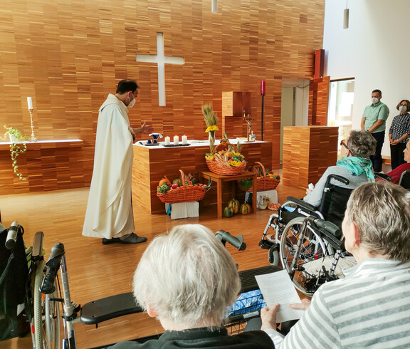 eine Erntedankmesse in einer Kirche. Ein Priester steht am Altar und hält den Gottesdienst, während Gemeindemitglieder, darunter ältere Menschen, in den Bänken sitzen und dem Geschehen folgen. Die Erntegaben sind festlich vor dem Altar arrangiert, und die Atmosphäre ist feierlich und besinnlich.