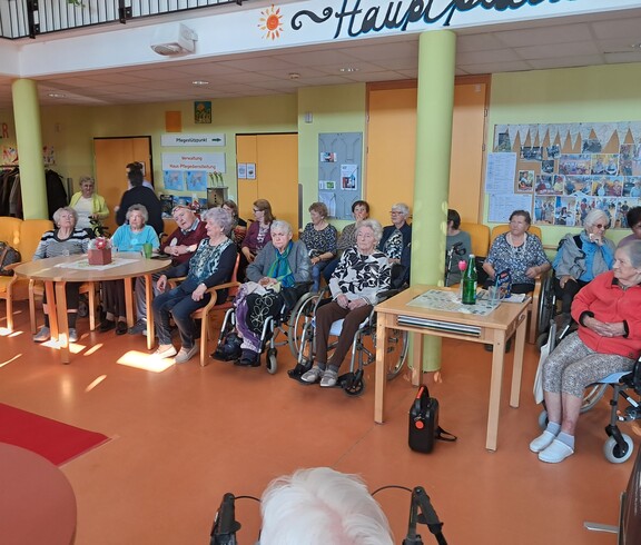 Bewohnerinnen und Bewohner des Pflegewohnhauses sitzen, teils mit Rollstühlen und Gehhilfen, im Gemeinschaftsbereich und verfolgen eine Modeschau.