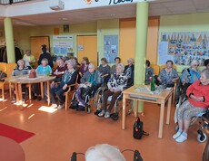 Bewohnerinnen und Bewohner des Pflegewohnhauses sitzen, teils mit Rollstühlen und Gehhilfen, im Gemeinschaftsbereich und verfolgen eine Modeschau.