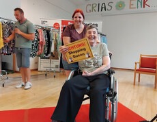 Bewohnerin im Rollstuhl hält Schild „Shopping Queen“, begleitet von Mitarbeiterin auf dem Laufsteg.
