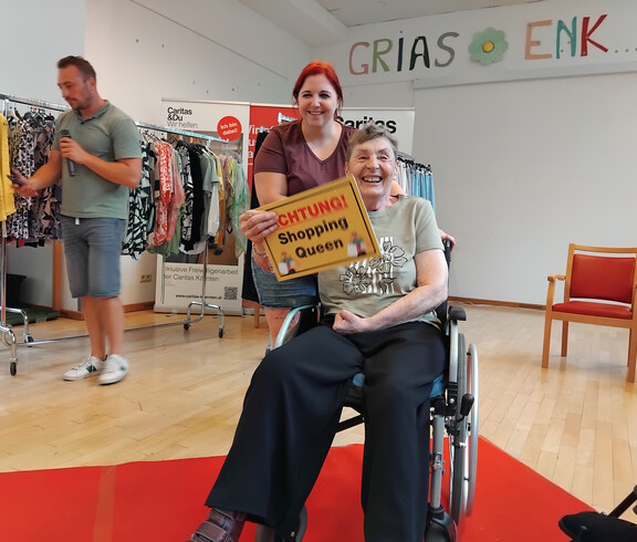 Bewohnerin im Rollstuhl hält Schild „Shopping Queen“, begleitet von Mitarbeiterin auf dem Laufsteg.