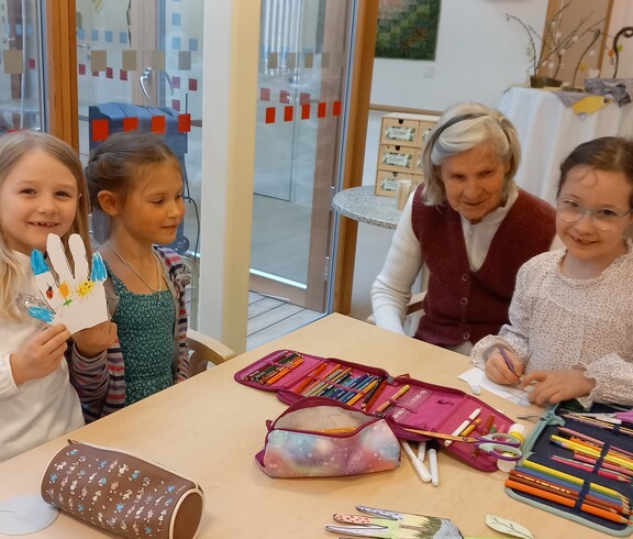 An einem Tisch im Gemeinschaftsraum des Pflegewohnhauses basteln mehrere Kinder gemeinsam mit einer älteren Bewohnerin, wobei Stifte, Papier und Bastelmaterialien ausgebreitet sind.