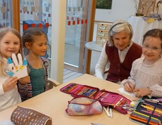 An einem Tisch im Gemeinschaftsraum des Pflegewohnhauses basteln mehrere Kinder gemeinsam mit einer älteren Bewohnerin, wobei Stifte, Papier und Bastelmaterialien ausgebreitet sind.
