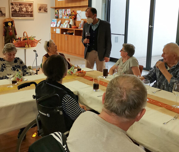 Eine Gruppe von Menschen, die an einem festlich gedeckten Tisch sitzen und sich unterhalten. Ein Mann steht und spricht mit den Gästen. Der Tisch ist herbstlich dekoriert, passend zum Erntedankfest. 