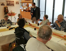 Eine Gruppe von Menschen, die an einem festlich gedeckten Tisch sitzen und sich unterhalten. Ein Mann steht und spricht mit den Gästen. Der Tisch ist herbstlich dekoriert, passend zum Erntedankfest. 