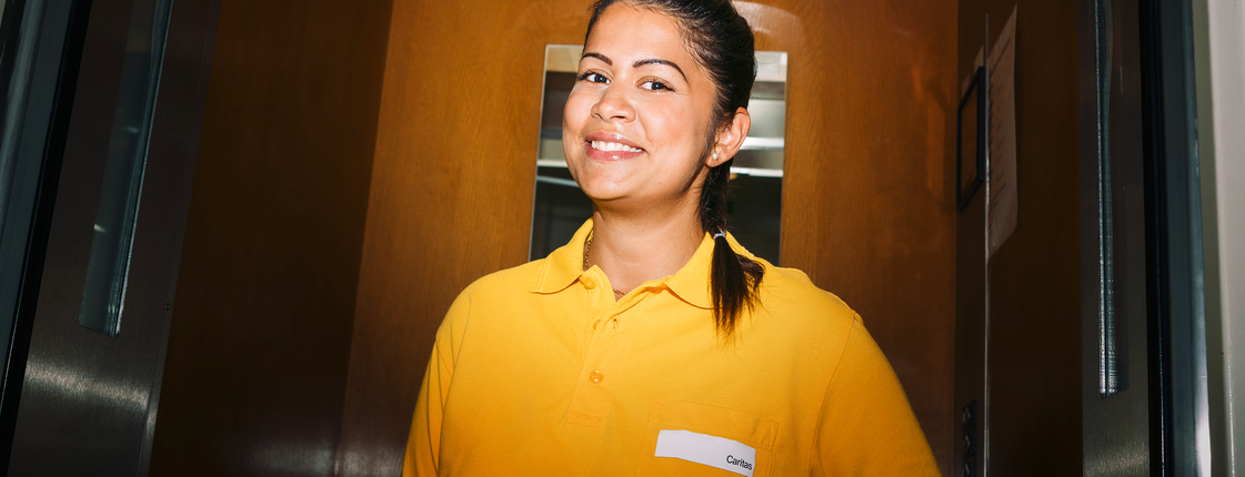 Eine junge Dame im orangenen Caritas-T-Shirt lächelt in die Kamera, sie steht in einem Aufzug.