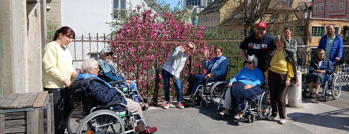 Menschen im Rollstuhl und Begleitpersonen stehen bei blühendem Strauch während eines gemeinsamen Spaziergangs durch Klagenfurt.