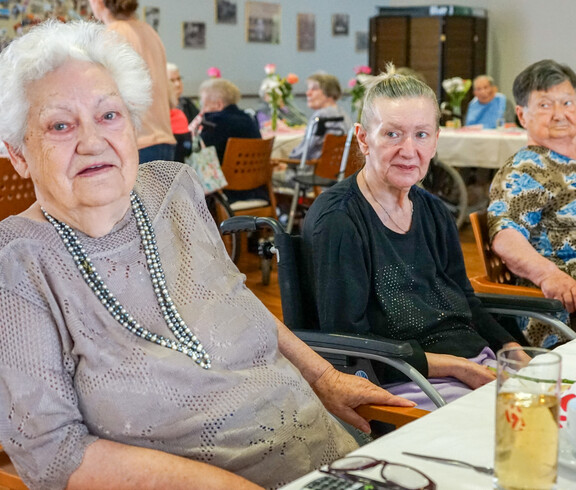 Drei Bewohnerinnen, die an einem Tisch bei der Muttertagsfeier sitzen. Sie wirken entspannt und genießen die Gesellschaft, während im Hintergrund weitere Gäste zusammenkommen.