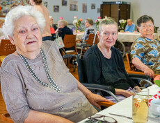 Drei Bewohnerinnen, die an einem Tisch bei der Muttertagsfeier sitzen. Sie wirken entspannt und genießen die Gesellschaft, während im Hintergrund weitere Gäste zusammenkommen.