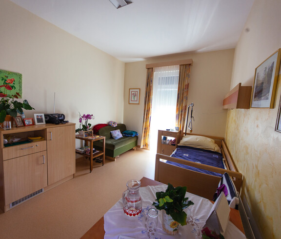 In diesem Raum befinden sich ein Bett, eine Couch, 2 Tische mit Blumen, ein Stuhl, 4 Bilder an der Wand, Fenster mit gelben Vorhängen sowie eine Kommode mit einem Radio, Bildern und einer Zimmerpflanze.