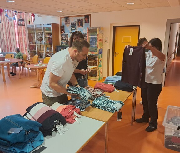 Mehrere Personen bereiten im Gemeinschaftsraum des Pflegewohnhauses Kleidung auf Tischen für eine Modeschau vor.