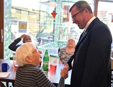 Eine Bewohnerin sitzt an einem Tisch im Aufenthaltsbereich des Pflegewohnhauses und begrüßt des Weihbischof im dunklen Anzug mit einem Handschlag, während weitere Bewohnerinnen im Hintergrund am Tisch sitzen.
