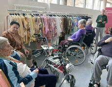 Gruppe von Bewohner*innen sitzt vor Kleiderständern mit farbiger Mode