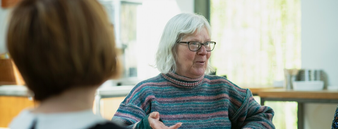 Bei einem Angehörigencafé erzählt eine Seniorin mit Brille, die eine Kaffeetasse in der Hand hält, den anderen Teilnehmerinnen von ihrem Alltag. 